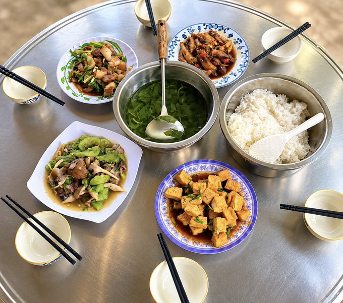 A Chinese home-cooked meal spread on a round metal table featuring a vegetable soup, white rice, tofu dish, stir-fried mushrooms with peppers, and other meat dishes.