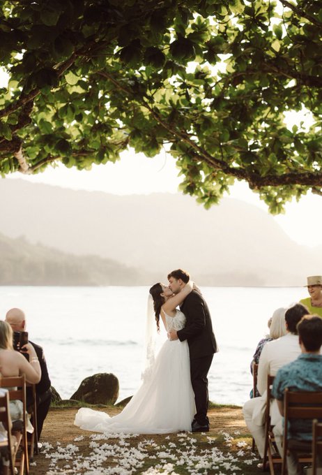 The image shows a couple kissing under a tree, with a group of people sitting around them at an outdoor wedding ceremony. The bride wears a white dress, while the groom is dressed in black. They are surrounded by chairs where guests have been seated throughout the event. The scene captures a moment of love and celebration as the newlyweds share their first kiss as husband and wife.