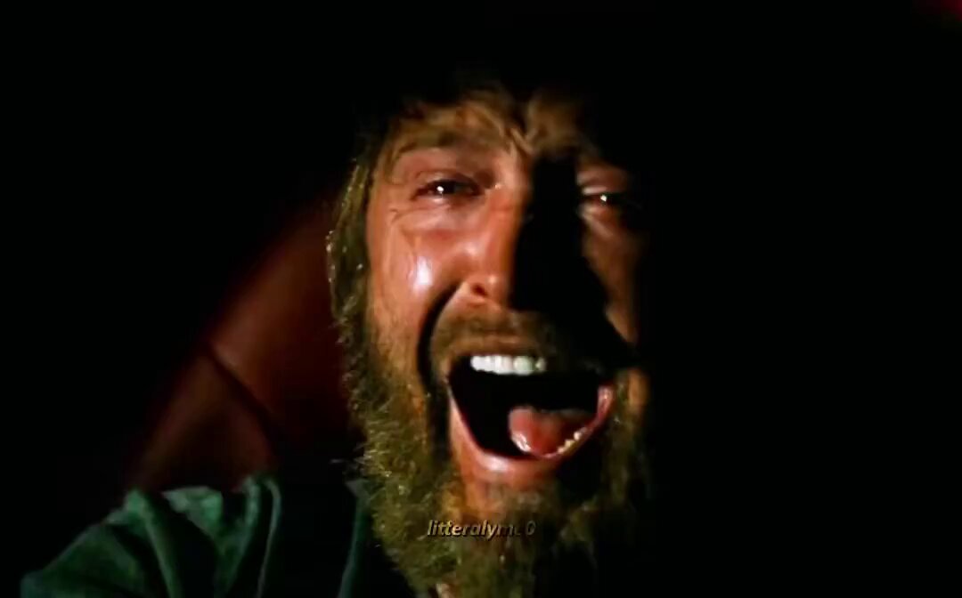 The image shows a man with a beard, wearing a green shirt, laughing heartily while sitting down. The background appears dark, which contrasts with his bright expression. There are no texts or other objects visible in the image, so we can only focus on the man and his reaction to something that has caught his attention.