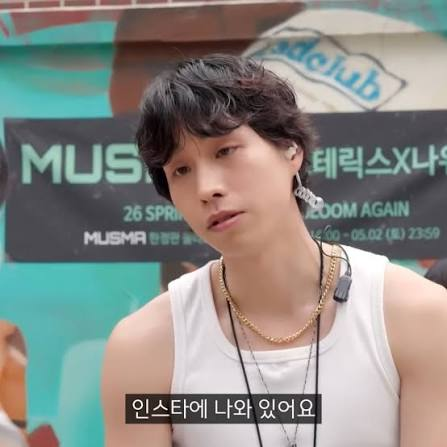 A young man with dark hair wearing a white tank top and gold chain, with Korean subtitles '인스타에 나와 있어요' visible at the bottom, appearing to be at a music event.