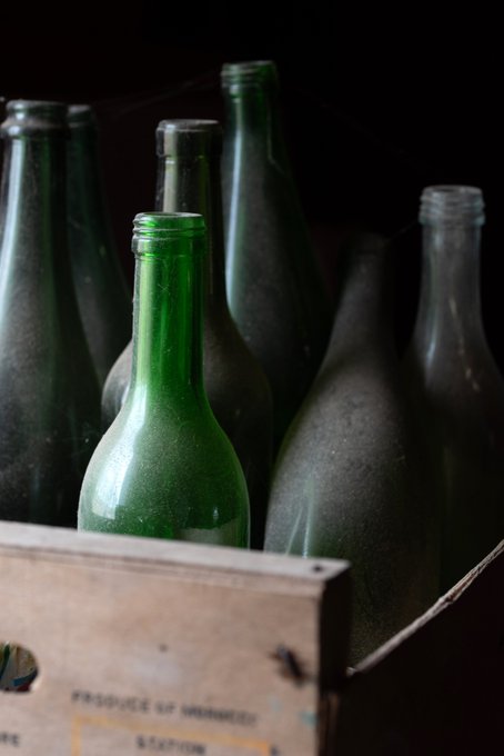 Several green glass bottles are clustered together in a wooden crate against a dark background, with partial text 'PRODUCE OF DOMINICA' and 'STATION' visible on the crate.