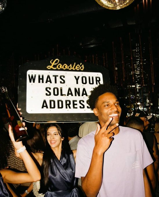 A smiling man in a purple shirt flashes a peace sign beside a woman in a black dress at what appears to be a nightclub, standing in front of a "Loosie's" sign reading "WHATS YOUR SOLANA ADDRESS" — capturing the playful moment referenced in the post about crypto traders eagerly waiting to hear that question.