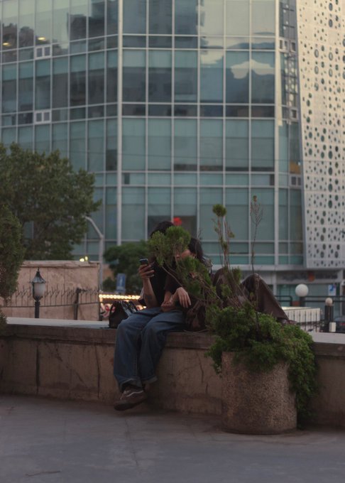 A person wearing a hooded jacket and jeans sits on a concrete ledge looking at their phone, with a large planter and glass skyscraper in the background.