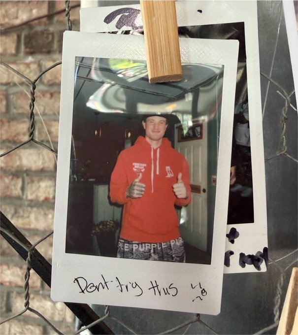 A polaroid photo clipped to a chain-link fence, showing a young man wearing a red hoodie and boxers giving two thumbs up; the caption 'Dont try this' is handwritten at the bottom.