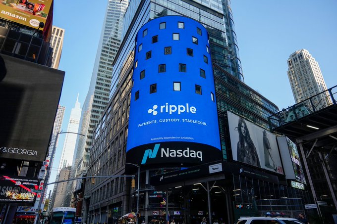 A large blue digital billboard in a busy city intersection displays the "ripple" logo with the tagline "PAYMENTS. CUSTODY. STABLECOIN" and a disclaimer reading "Availability dependent on jurisdiction," positioned above the Nasdaq logo. The image directly supports the post by showing Ripple's branding featured on the iconic Nasdaq tower, highlighting a notable connection between the XRP/XRPL ecosystem and the Nasdaq stock exchange.