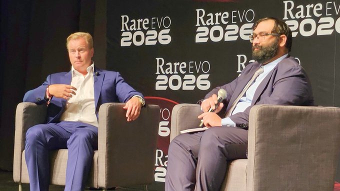 Two men in suits sit in armchairs on a stage at the 'RareEVO 2026' conference, with one man holding a microphone.