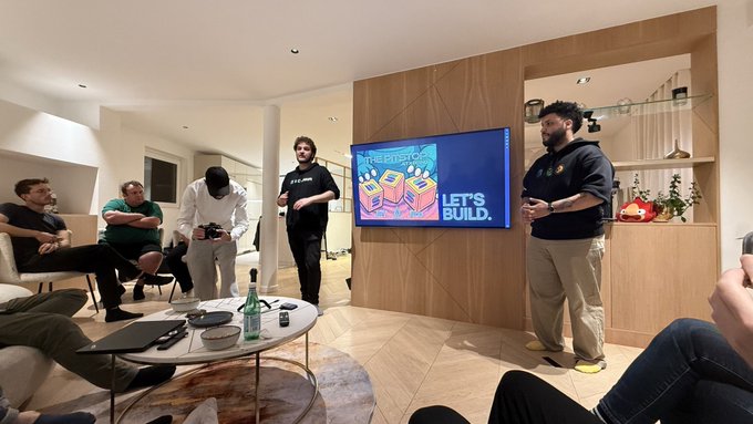A man in a black hoodie stands presenting in a living room in front of a TV displaying 'BREADTOWN LET'S BUILD', while a group sits around a coffee table.