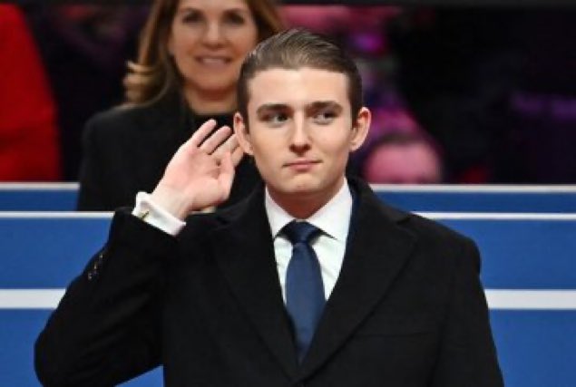 A young man in a dark suit with a navy tie being sworn in with his right hand raised, while a woman stands behind him, appearing to be at a formal ceremony or inauguration.