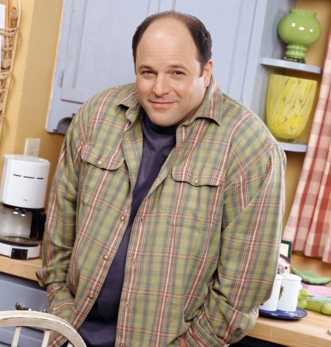 Photo of actor Jason Alexander as George Costanza from Seinfeld, wearing a green plaid shirt over a dark t-shirt, standing in a kitchen next to a coffee maker and a green lamp.