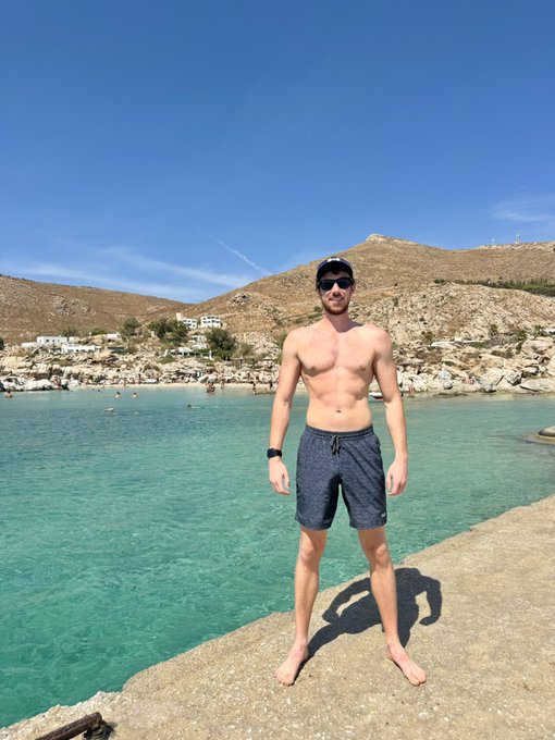 A shirtless man in sunglasses, a cap, and dark swim trunks stands on a rocky beach by clear turquoise water, with arid hills and a small village in the background.