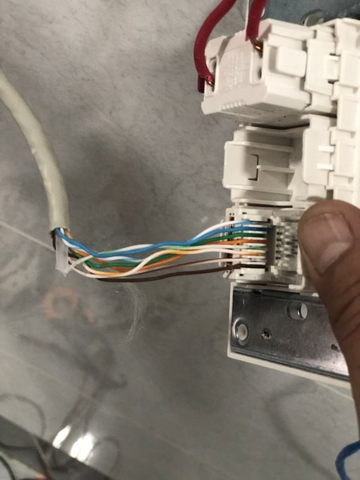 Close-up photo of electrical wiring with multiple colored wires (blue, orange, green, brown) being held by a person's thumb, showing the internal components of what appears to be a white electrical junction box or outlet.