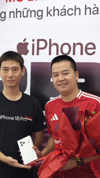 Two men posing at an iPhone promotional event; one wears a black shirt while the other in a red jersey holds a bouquet of red roses and a bouquet wrapped in red paper, displaying an iPhone 18 Pro phone in front of an 'iPhone' branded backdrop.
