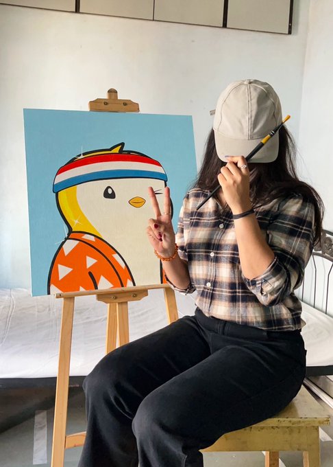 A young woman wearing a beige cap and plaid shirt, holding a paintbrush to her face while making a peace sign, sitting next to a canvas with a painting of a duck-like character in athletic wear.