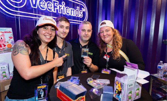 Four people smiling behind a table at a convention, with a neon 'VeeFriends' sign in the background and various collectible boxes and graded cards displayed on the table.