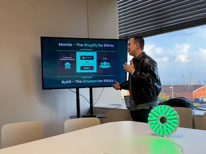 A man in a black leather jacket gives a presentation in a modern conference room, gesturing toward a screen displaying a slide titled "Mantle - The Shopify for RWAs" which outlines asset distribution between traditional institutions and retail users, while a green 3D-printed fan model sits on the table in the foreground. The image captures the moment from one year ago when the RWAccelerate vision was first introduced, highlighting Mantle's positioning as the Shopify for real-world assets that the post reflects on.