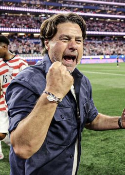 A man in a dark blue shirt and a wristwatch celebrates enthusiastically with a clenched fist and an open mouth on the sidelines of a soccer stadium.