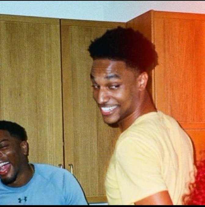 A young man with a faded haircut smiles while wearing a pale yellow t-shirt, standing next to another person laughing in a gray Under Armour shirt, with wooden cabinets visible in the background. The image captures a casual, joyful moment that matches the post's playful "current mood" caption referencing $ANSEM.