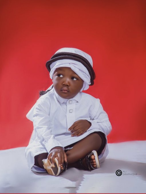 A studio portrait of a young child dressed in traditional white Emirati attire with a white and black headdress, sitting against a red backdrop, with 'ALI GRAPHIQUE' watermark visible.