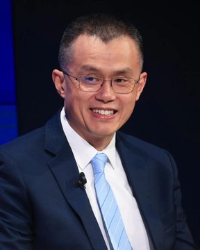 A man wearing glasses, a dark suit, white shirt, and light blue tie smiles while speaking at an event, with a small microphone clipped to his lapel. The image complements the quote by Binance founder Changpeng Zhao (CZ), emphasizing his message of focusing on building rather than reacting to distractions in the crypto industry.