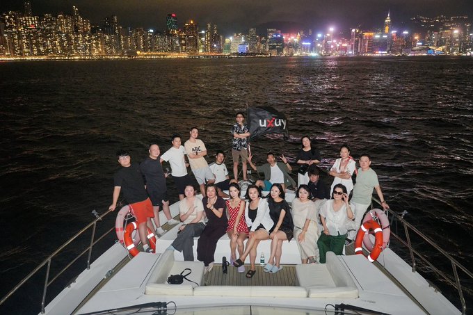 A group of people posing on the bow of a yacht at night with the Hong Kong skyline illuminated in the background and a 'LUXU' flag held aloft.