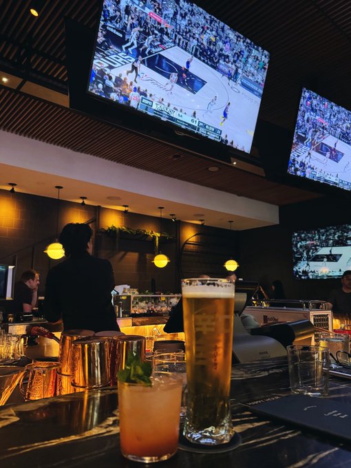 The image shows a bar scene with two glasses of beer, one tall glass filled with beer and another smaller glass containing a cocktail. A television screen displays a basketball game, adding to the lively atmosphere. The presence of people at the bar suggests that they are enjoying their time watching sports or engaging in conversation while sipping on their drinks.