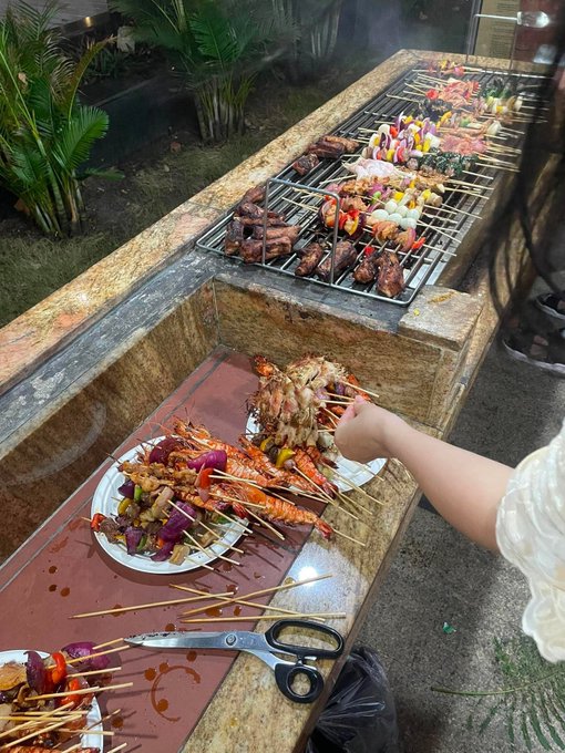 An outdoor barbecue scene featuring a long grill filled with skewers of meat, shrimp, vegetables, and peppers, with a person's hand reaching in to grab food and a pair of scissors resting on plates of prepared skewers in the foreground.