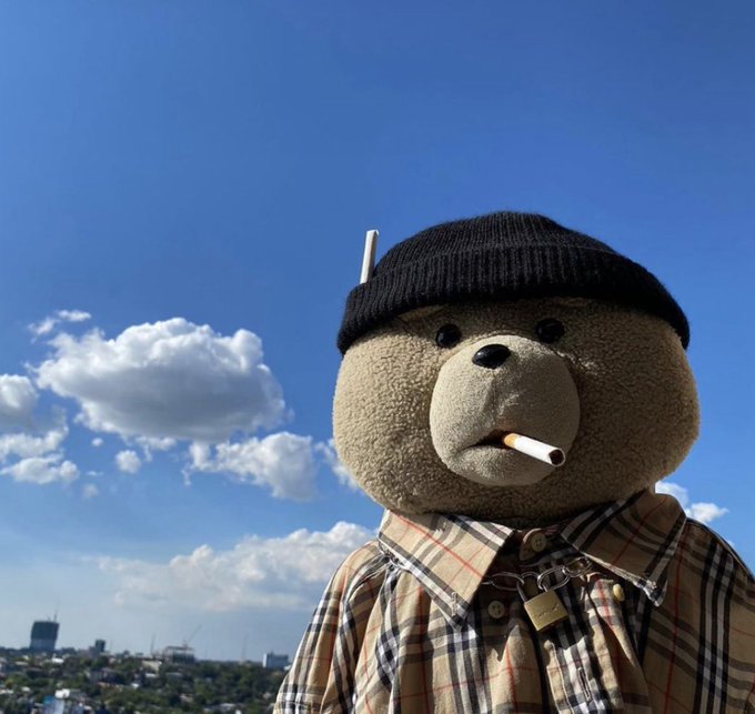 A teddy bear wearing a black beanie and a plaid shirt with a padlock necklace has a cigarette in its mouth, set against a blue sky with clouds.