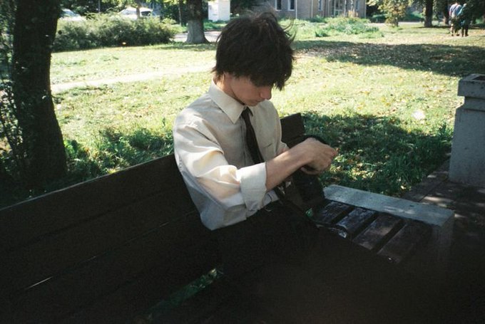 A young man with dark hair sits on a wooden park bench, wearing a white dress shirt and black tie, looking down as he adjusts or examines something in his hands—perhaps a camera or device—on a sunny grassy campus. The image complements the post's "touch grass" weekend reminder by showing someone taking a break outdoors, though still preoccupied with a gadget rather than fully disconnecting.