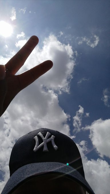 A low-angle selfie showing a person making a peace sign against a bright blue sky with scattered clouds, wearing a navy blue New York Yankees cap visible at the bottom of the frame.