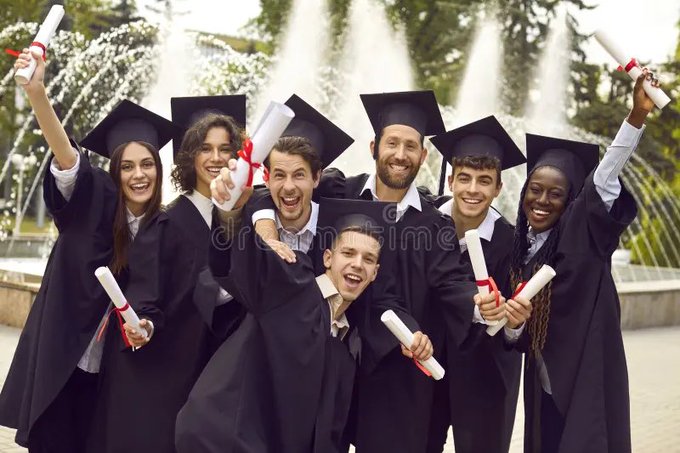 Group of seven graduates in black caps and gowns cheering and holding up diplomas in front of a fountain.