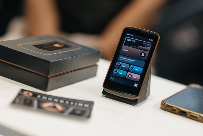 A smartphone displaying a Bitcoin wallet interface showing a balance of $64,178, placed on a display stand next to its packaging.