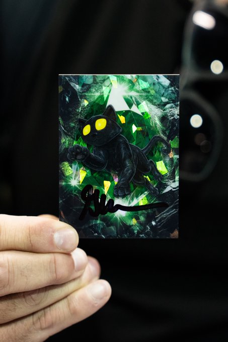 A hand holds up a holographic trading card featuring a black cat with glowing yellow eyes against a shimmering green crystalline background, with a signature visible at the bottom.
