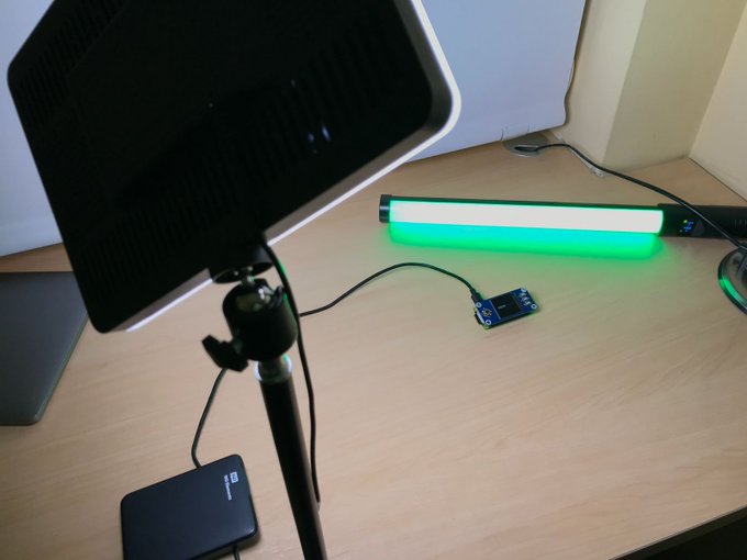 A workspace setup on a wooden desk featuring a large black softbox light on a stand and a green LED light bar connected to a small device.