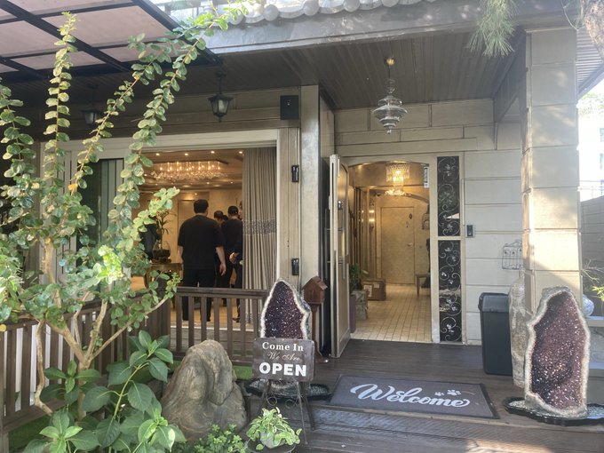 The entrance of a cozy establishment with a 'Come In We're Open' chalkboard sign, an 'OPEN' sign, and a 'Welcome' doormat, decorated with greenery and large geode stones.