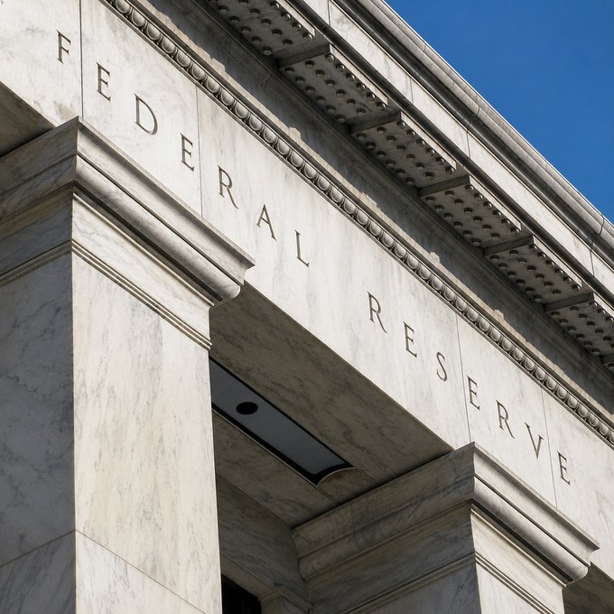 A low-angle view of the marble façade of a Federal Reserve building, with the engraved words "FEDERAL RESERVE" prominently displayed above the entrance against a clear blue sky. The classical architecture symbolizes the institution expected to raise interest rates by 25 basis points.