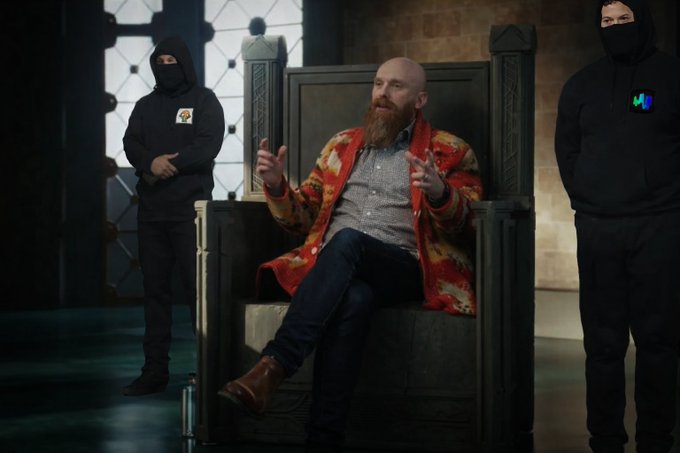 A bearded man in a red and yellow patterned jacket sits on a large throne-like chair gesturing with his hands, flanked by two figures in black hoodies and face masks.