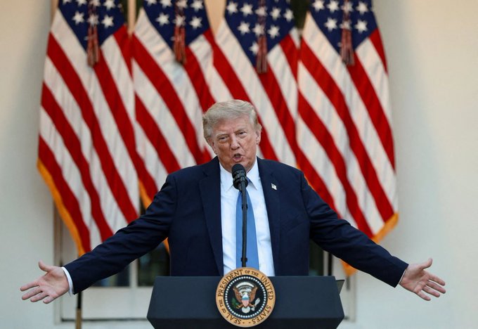 Former US President Donald Trump speaking at a podium bearing the Presidential Seal, standing in front of multiple American flags with his arms outstretched.