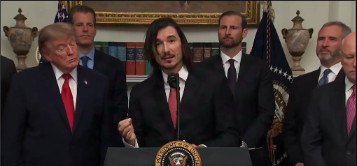 A man with long dark hair and a beard speaks into a podium bearing the seal of the President of the United States, flanked by Donald Trump on the left and several other men in suits, with American flags and a formal room in the background. The image appears to depict a White House event, though it does not directly illustrate the post's rumor about Trump launching a token on Robinhood.