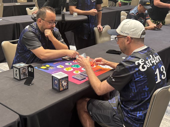 Photo of two men playing a trading card game at a tournament table, with one wearing a shirt that says 'Cst4lfn 13'.