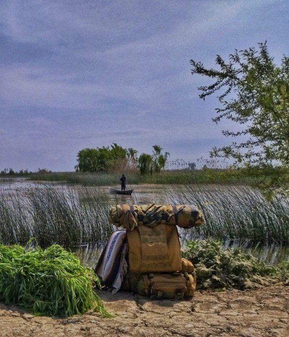 A scenic view of a wetland or lake with a small boat in the distance, featuring a tan tactical backpack and a bag of greens sitting on the muddy shore in the foreground.