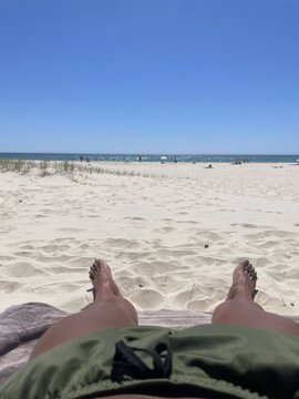 A first-person view of someone relaxing on a sandy beach, with their bare feet extended toward the ocean under a clear blue sky.