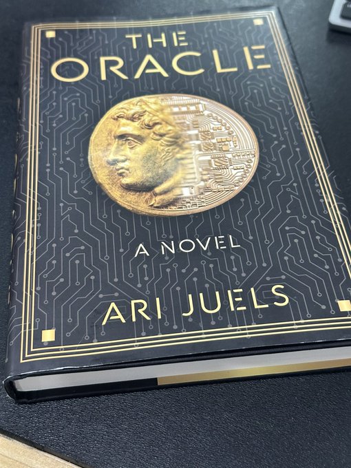 A hardcover copy of "The Oracle: A Novel" by Ari Juels rests on a dark surface, featuring a striking cover with a classical gold medallion profile merged with circuit board patterns against a black background. The book's title and tech-inspired design playfully tie into the post's theme about oracles in the crypto world, referencing the dual meaning of "oracle" as both prophecy and financial infrastructure ($LINK/Chainlink).