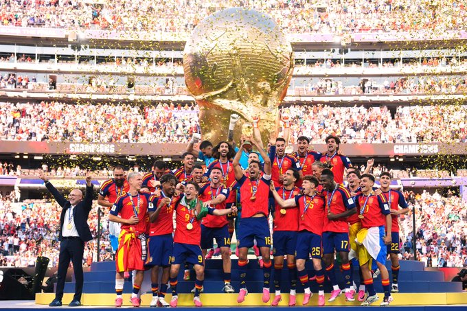 The Spanish national football team celebrating on a podium while lifting the FIFA World Cup trophy in a packed stadium, with players wearing red jerseys and gold medals around their necks.