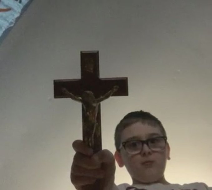 A young boy with glasses holds up a crucifix featuring Jesus against a plain wall.