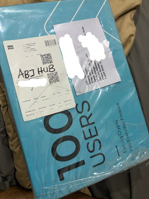 A sealed blue Bitget Wallet box labeled "100 USERS - Turning Crypto Finance into Everyday Finance" with an "ABJ HUB" shipping label rests on a bed, matching the creator's teaser of an upcoming Bitget Wallet unboxing reveal.