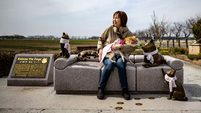 A woman sits on a stone bench surrounded by multiple dog statues wearing scarves, at what appears to be the "Kabosu the Doge" memorial site.