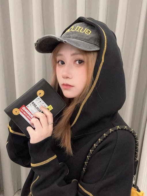 A young woman wearing a black hoodie and a faded black cap poses with a black notebook wallet featuring colorful stickers.