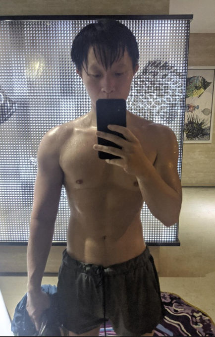 A lean, shirtless man takes a mirror selfie in what appears to be a hotel room, showcasing his toned physique as a testament to natural fitness at age 49. The caption celebrates this as a real, unedited body achieved through hard work, promoting health as a more valuable pursuit than material wealth.