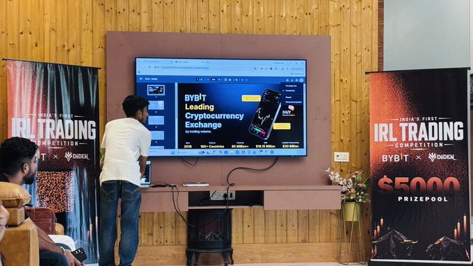 A man interacts with a large screen displaying the ByBit cryptocurrency exchange, flanked by banners advertising 'India's First IRL Trading Competition' with a $5000 prizepool, sponsored by ByBit x CoinDCX.
