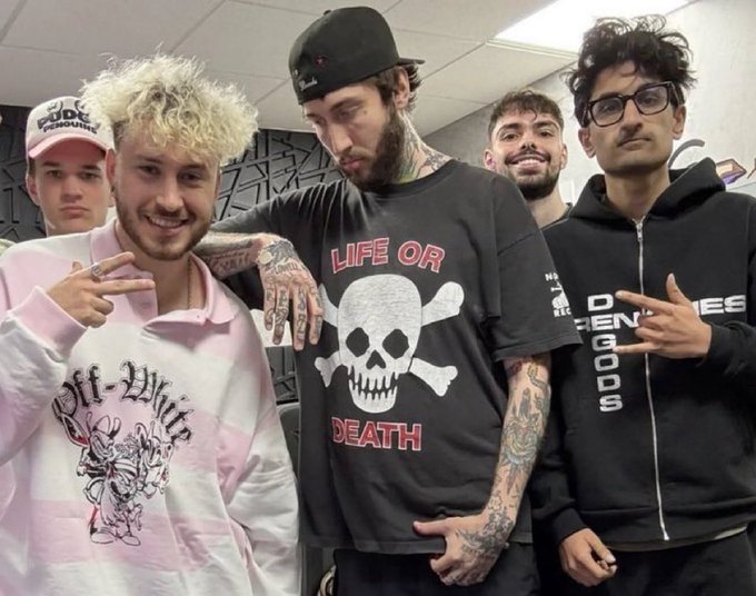 Four young men pose together in an indoor setting; the central figure has a heavily tattooed neck and arms, and wears a black t-shirt with a skull and crossbones graphic reading 'LIFE OR DEATH'.